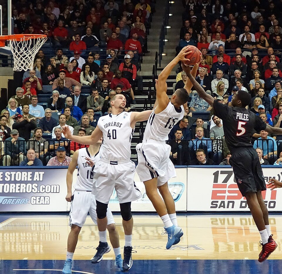 Basketball game with a Toreros player in white driving past a defender in black.