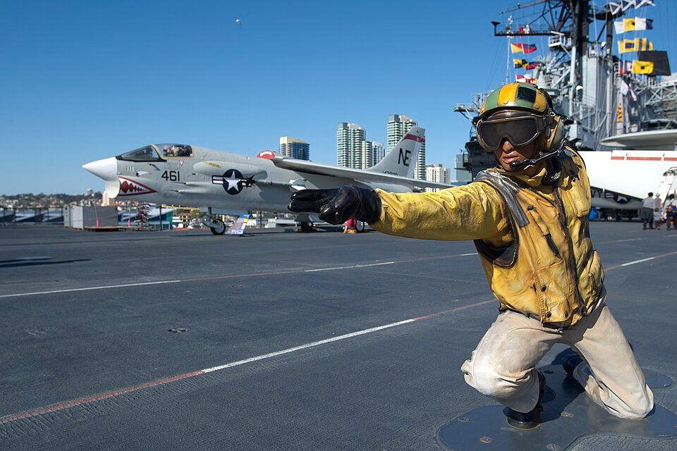 A sailor figure on the flight deck of the USS Midway signaling an aircraft, with a jet behind