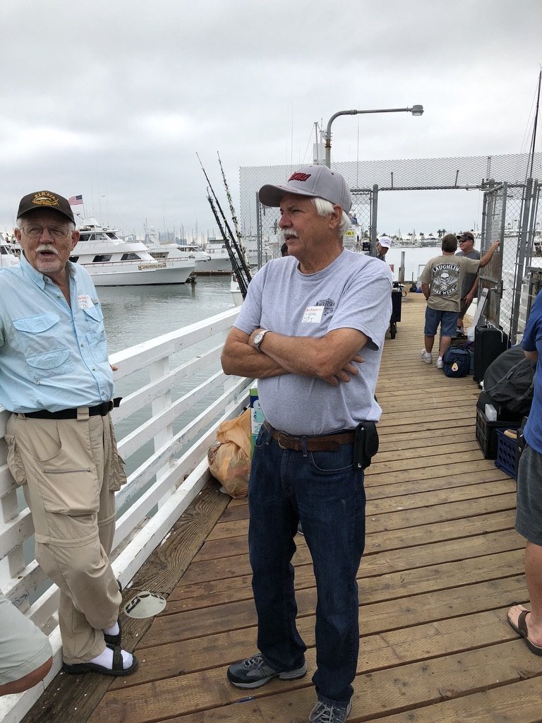 Two anglers talking on a wooden dock before a trip, boats and rods behind them