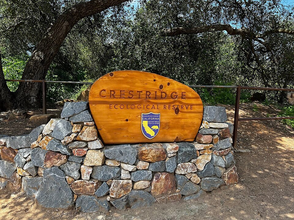 A carved wooden sign reading Crestridge Ecological Reserve mounted on a stone wall under oak trees.
