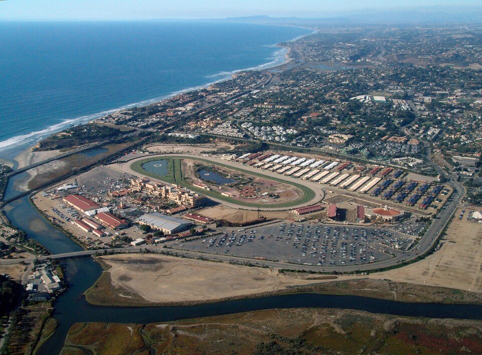 An aerial view of the Del Mar Fairgrounds beside the coast and lagoon, with its buildings, track and parking lots