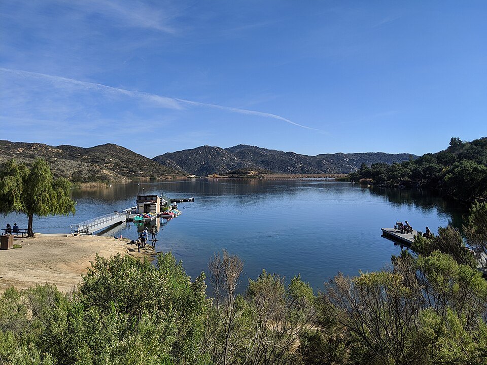 A calm lake ringed by brushy hills with a fishing dock and a few people on the shore.