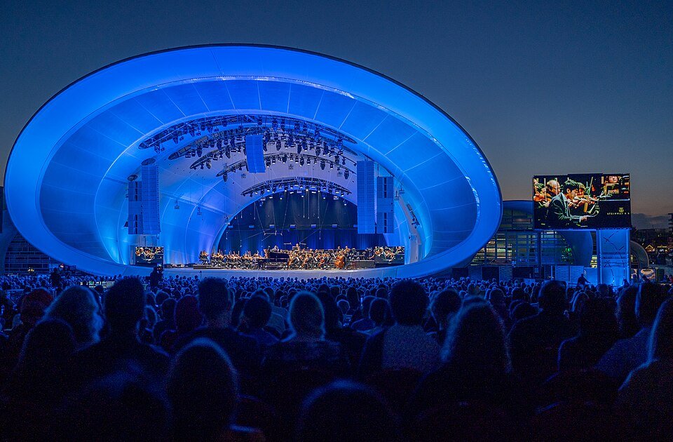 Concert crowd at dusk facing a lit blue band shell stage with an orchestra on it.