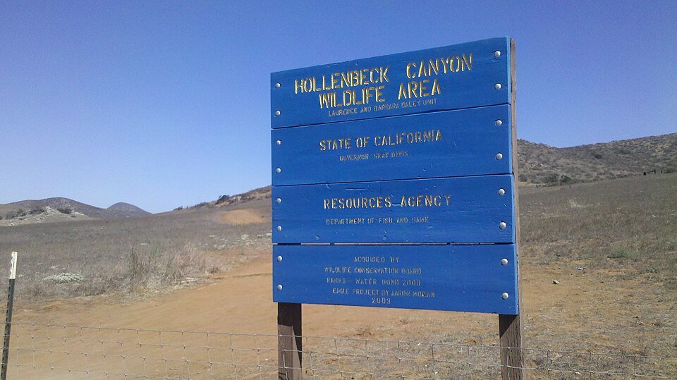 The blue Hollenbeck Canyon Wildlife Area entrance sign on a dry hillside