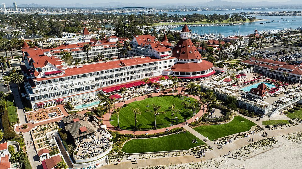An aerial view of the Hotel del Coronado with its red roofs and turret, the green lawn and the beach