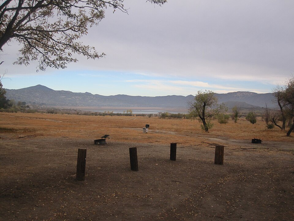 Dry campground with wooden posts and fire rings in front of a lake and mountains.