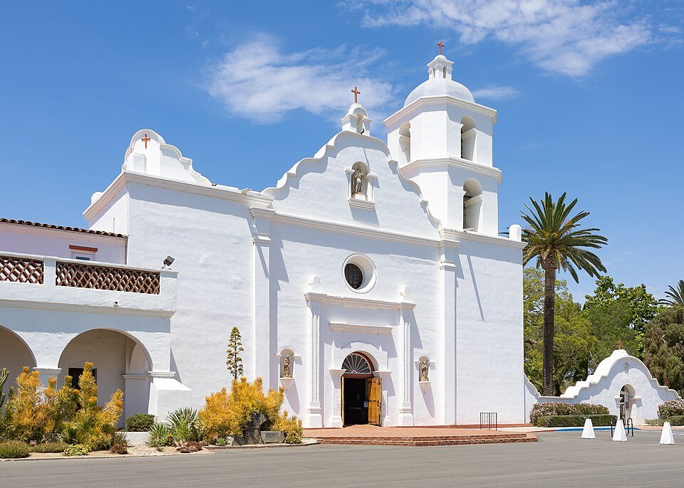 The white front of Mission San Luis Rey with its bell tower and a palm tree under a blue sky