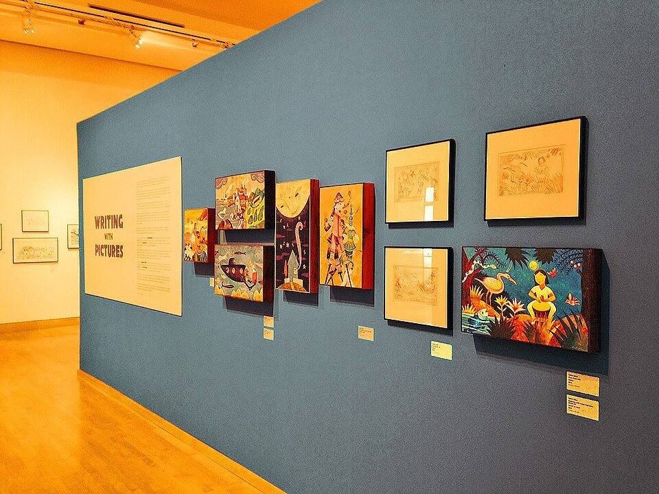 Framed storybook art on a blue gallery wall in an exhibit at the California Center for the Arts, Escondido