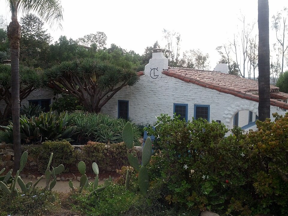 White adobe ranch buildings with a red tile roof among cactus and palm trees.