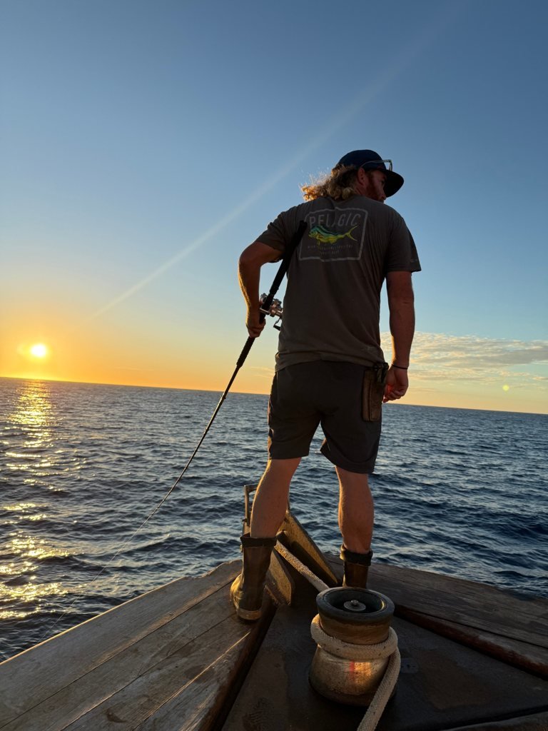 A deckhand on the bow at sunset holding a rod, the sun low over a calm ocean