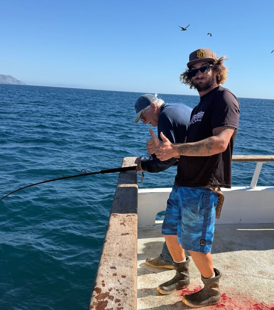 A crew member on deck giving a thumbs up beside an angler working a bent rod at the rail over blue water