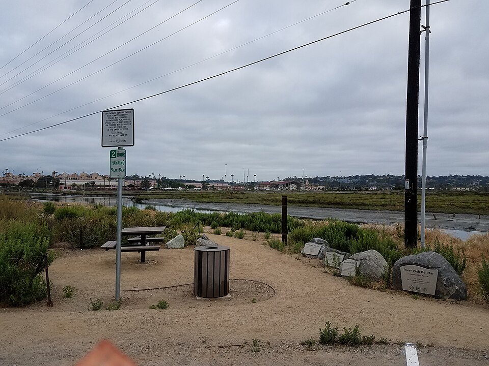 Dirt path and picnic table beside a tidal wetland, with a sign post and cloudy sky