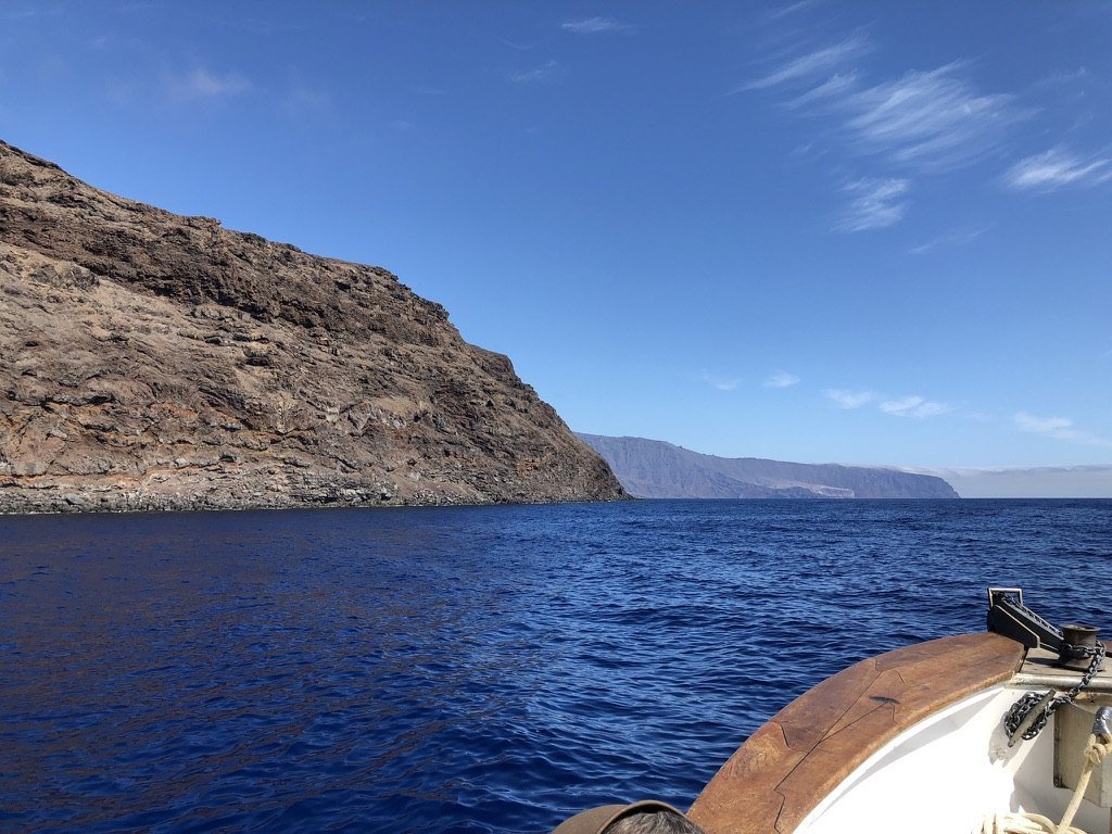 A tall brown rock cliff rising from deep blue water under a clear sky