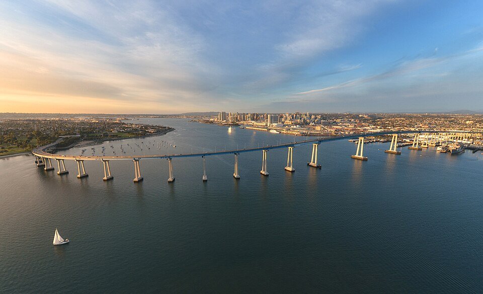 An aerial view of San Diego Bay at golden hour, with the Coronado Bridge crossing the water and a sailboat below