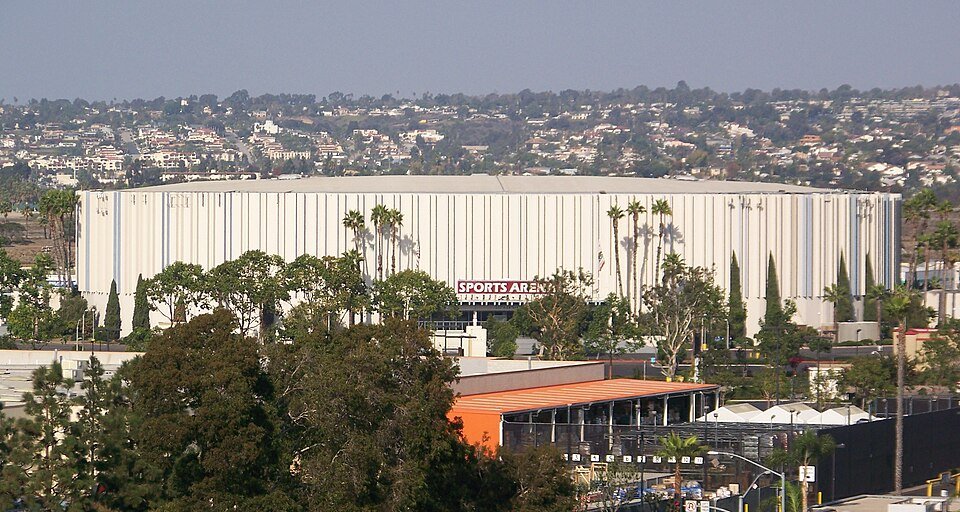 A large cream-colored arena building with palm trees and homes on the hill behind it