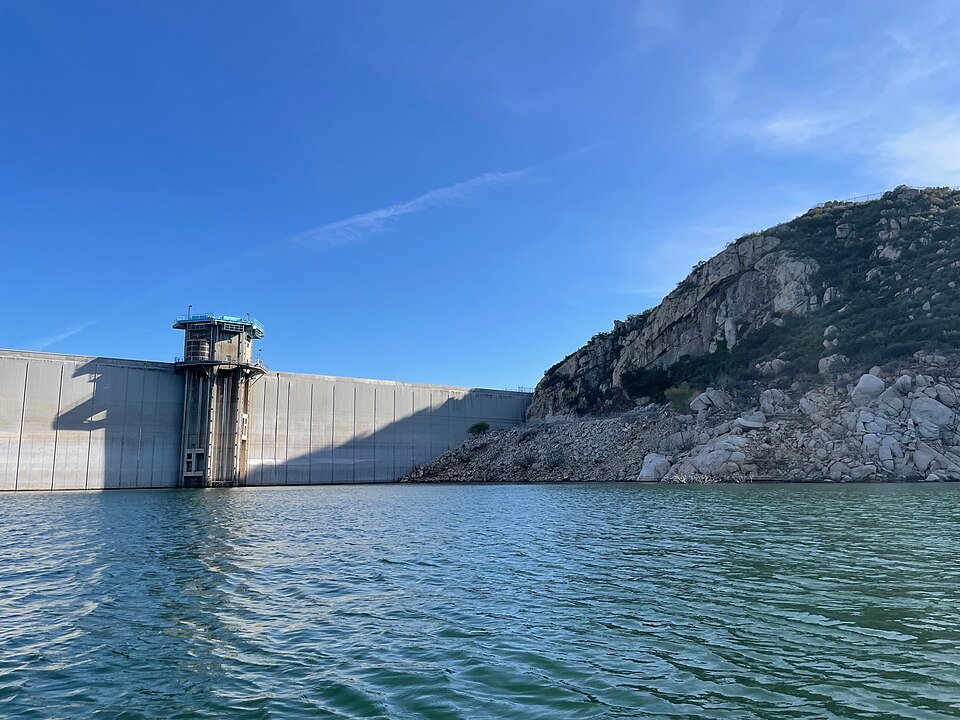The concrete San Vicente Dam and its intake tower beside a rocky hillside, seen from the water