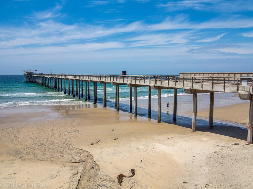Long concrete pier extending over a sandy beach and surf under blue sky