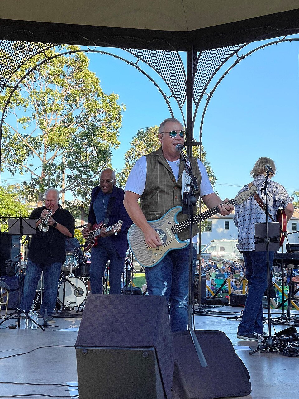 A guitarist singing with a small band playing under a gazebo-style bandstand on a sunny day, with trees behind.
