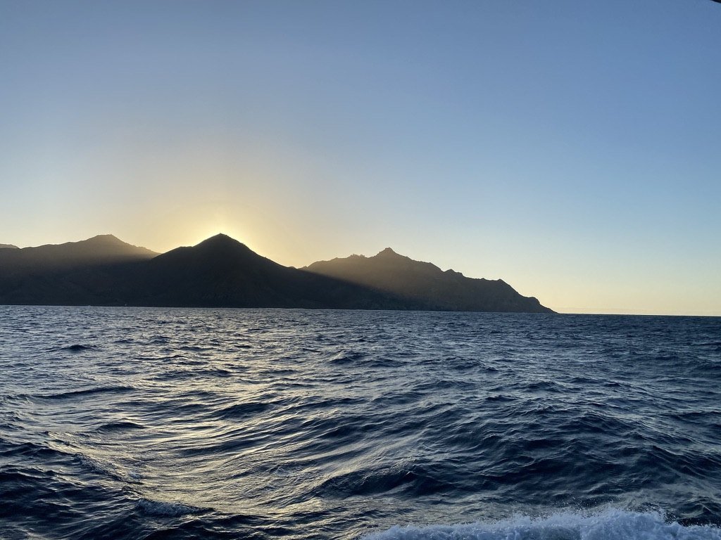 Sun rising behind a dark island ridge over deep blue ocean, seen from a boat
