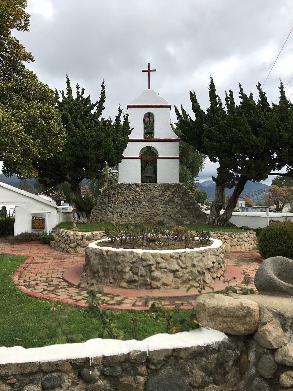 A white mission bell tower with a cross above a stone-ringed well and garden, surrounded by trees