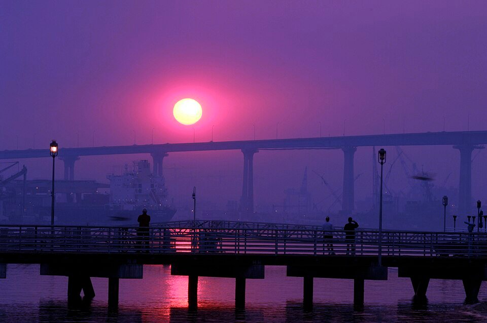 People on the Coronado ferry landing pier at sunrise, with the Coronado Bridge behind and a glowing sun