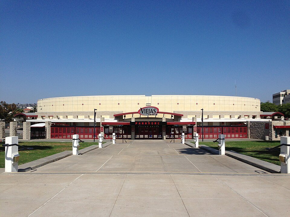 Round tan arena building with red entrance doors and sign under a deep blue sky
