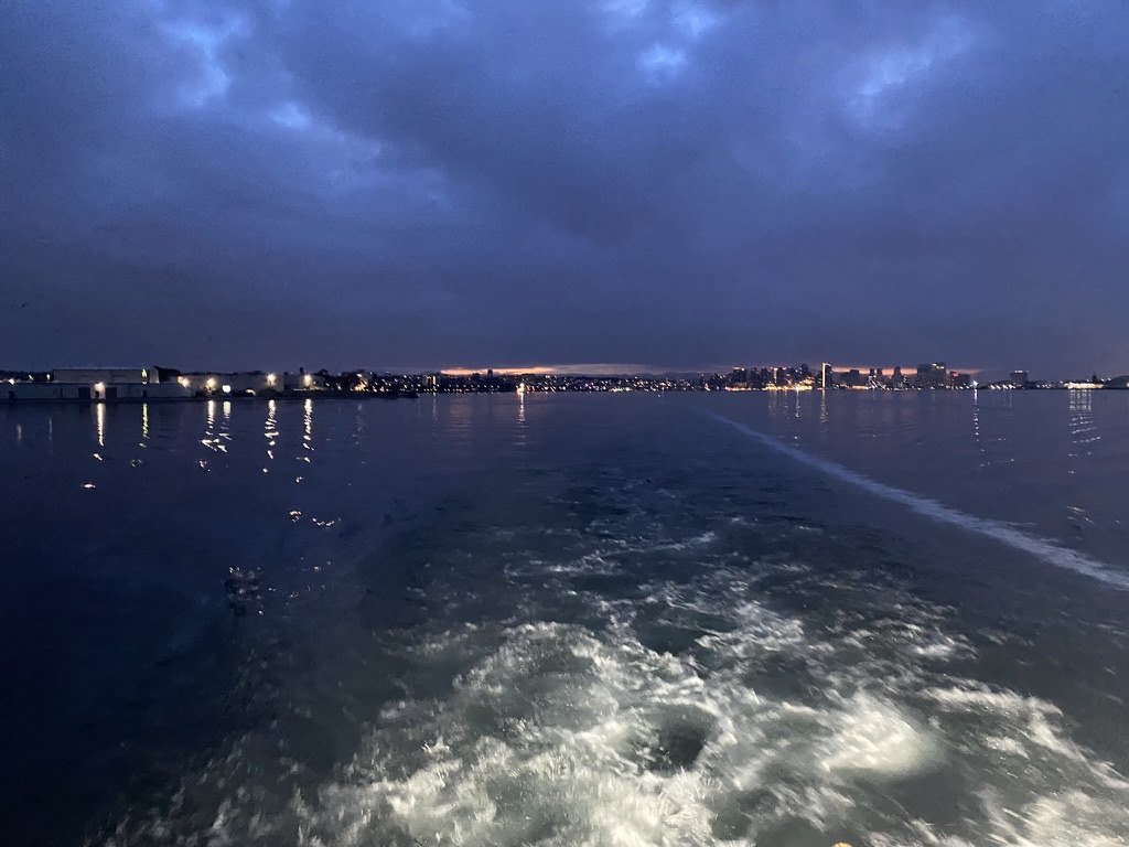Churned white wake on dark water with distant shore lights at night