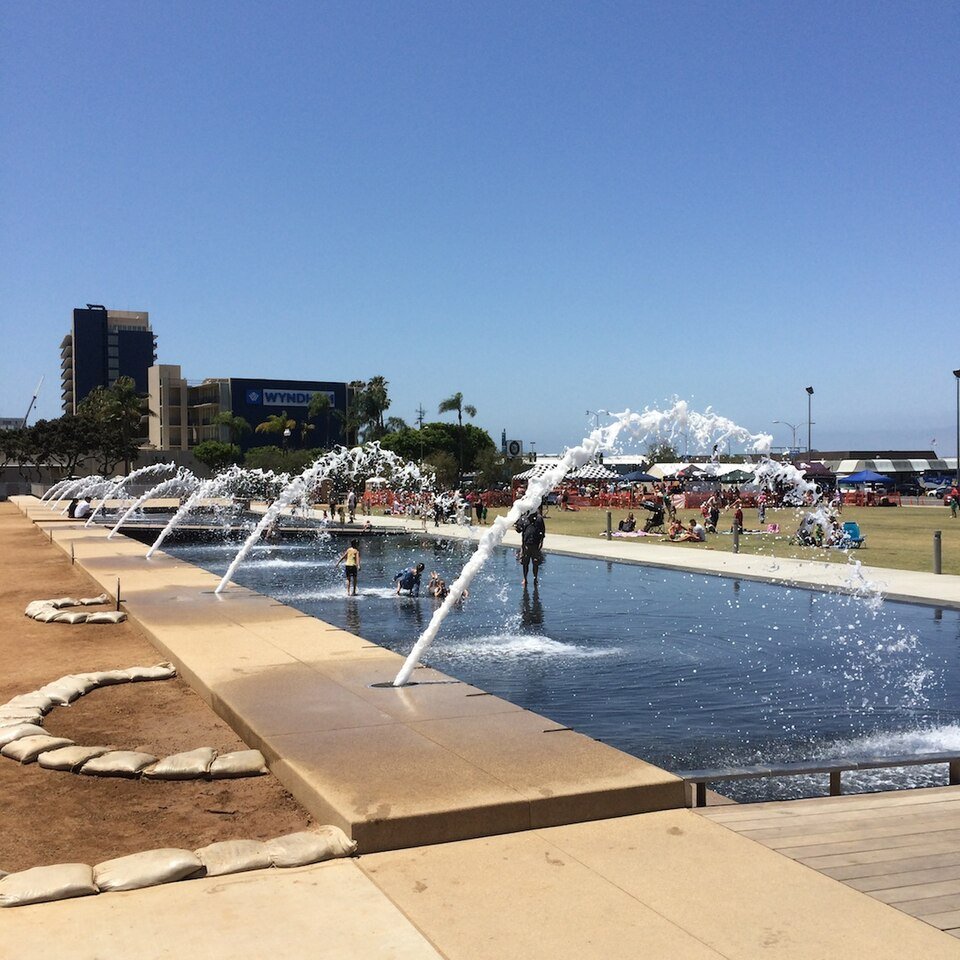 Fountains playing along a long shallow pool, with people on the lawn beyond and buildings behind.
